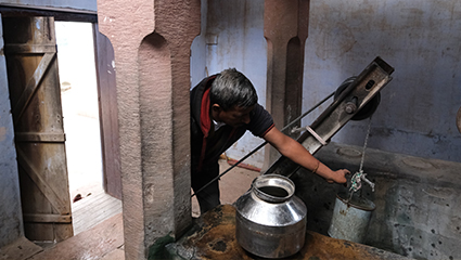 जैता बेरा से स्टेनलेस स्टील के घड़े (पानी के बर्तन) में अपने घर पानी ले जाता एक व्यक्ति। असलम सैय्यद जल झरोखा वर्कशॉप द्वारा फोटो दिसंबर २०२१